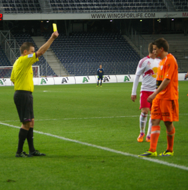 Ein Schiedsrichter zeigt einem Fußballspieler auf dem Feld eine gelbe Karte, umgeben von einer Gruppe von Menschen, mit einem Torpfosten, einem Zaun, einer Tafel, Metallrahmen, einem Bildschirm, Stühlen und einer Decke mit Deckenleuchten im Hintergrund.