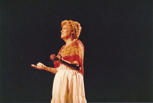 Eine Frau in einem weißen, hochgeschlossenen, langärmligen Kleid steht auf einer dunklen Bühne mit ausgestreckten Armen, ihr Haar ist zurückgebunden und ihr Gesichtsausdruck ernst.