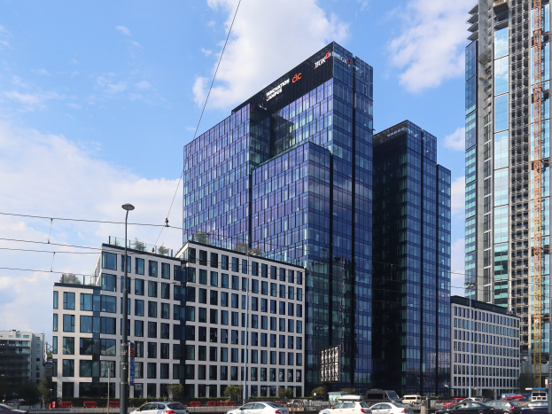 Ein großes Gebäude mit vielen Fenstern, das als Hauptsitz der Deutschen Bank identifiziert wurde, steht in der Mitte einer Stadt umgeben von Straßenlaternen, Strommasten, Stromkabeln, Kraftfahrzeugen, Büschen, Bäumen und einem bewölkten Himmel.