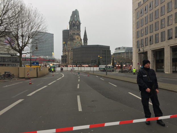 Ein Polizist in schwarzer Uniform und Mütze steht auf einer Straße in Berlin umgeben von Absperrgittern, Fahrrädern, Laternen, Bäumen und Gebäuden, mit einem Uhrenturm und einem klaren blauen Himmel im Hintergrund.