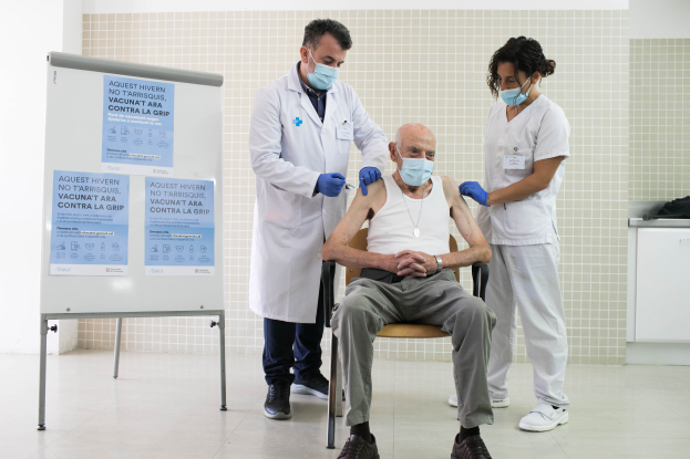 Ein älterer Mann sitzt in einem Stuhl und erhält von zwei maskierten und handschuhgetragenen Ärzten in einer Krankenhausumgebung eine Impfung. Links befindet sich eine Tafel und rechts einige Gegenstände auf einer Plattform.