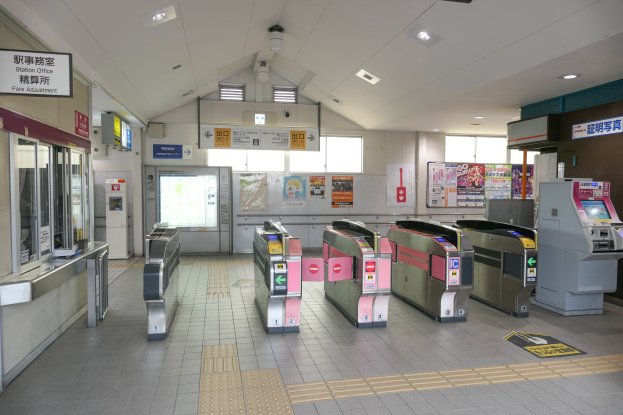 Ein Bahnhof mit einer Reihe von Metalldetektoren, eine Plattform mit Glastüren und Texttafeln links und Plakate an der Wand mit Deckenleuchten im Hintergrund.