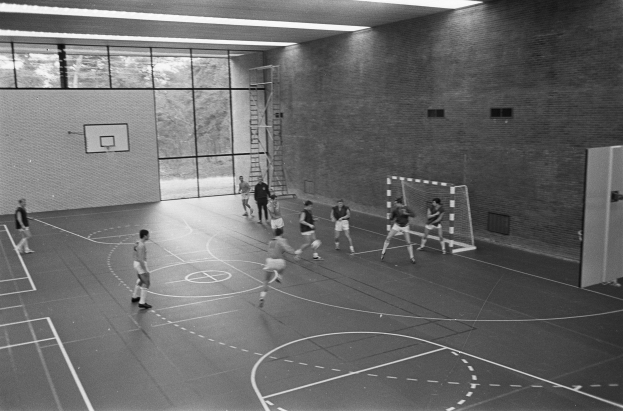 Ein Schwarz-Weiß-Bild einer Gruppe von Menschen, die Handball in einer Turnhalle spielen, mit sichtbaren Spielern, einem Torpfosten, einer Leiter, einer Wand, Fenstern und einer Decke mit Leuchten.