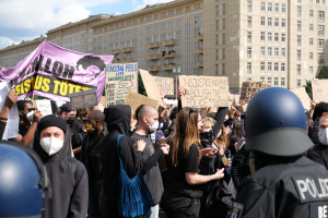 Eine Gruppe von Menschen mit Masken und Schildern protestiert vor einem Gebäude, wobei zwei Polizisten mit Helmen auf der rechten Seite zu sehen sind, vor einem Hintergrund aus Bäumen und bewölktem Himmel.