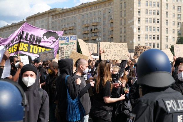 Eine Gruppe von Menschen mit Masken und Schildern protestiert vor einem Gebäude, wobei zwei Polizisten mit Helmen auf der rechten Seite zu sehen sind, vor einem Hintergrund aus Bäumen und bewölktem Himmel.