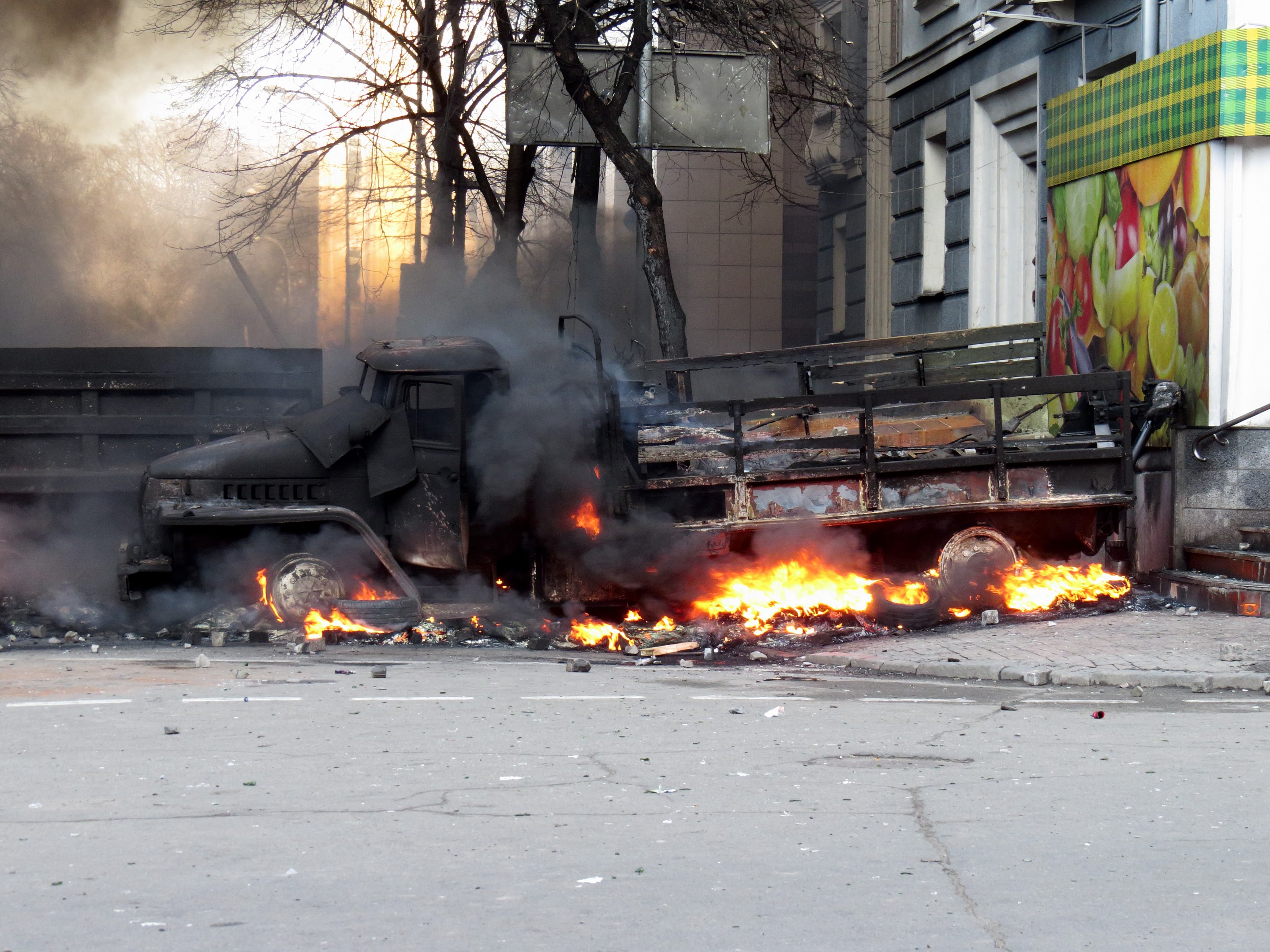 Ein Lkw brennt am Straßenrand, mit Rauch, der daraus aufsteigt, umgeben von Bäumen, Gebäuden und einem klaren Himmel.