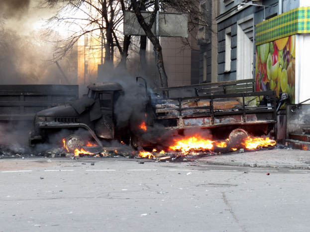 Ein Lkw brennt am Straßenrand, mit Rauch, der daraus aufsteigt, umgeben von Bäumen, Gebäuden und einem klaren Himmel.
