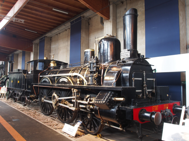 Eine große, alte Dampflokomotive, die in einem Museum ausgestellt ist, umgeben von Eisenbahnschienen, Informationsschildern und anderen Ausstellungsstücken, mit einer Wand und einer beleuchteten Decke im Hintergrund.