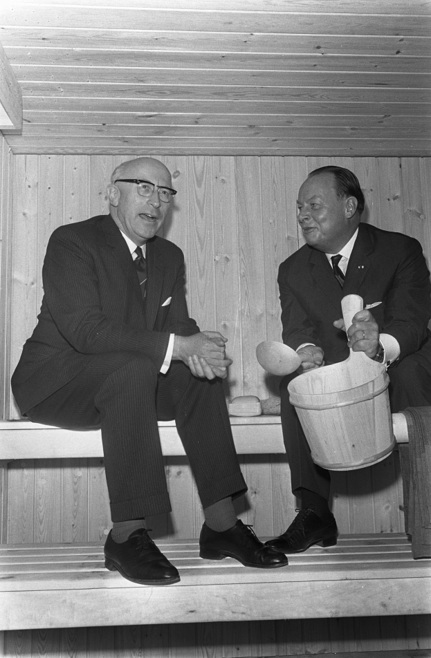 Zwei Männer in formeller Kleidung sitzen auf einer Holzbank in einer Sauna, einer hält einen Eimer, mit einem Tuch über der Bank, vor einer Holzwand.