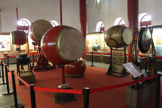 Ein Museumsraum mit verschiedenen Musikinstrumenten, darunter Trommeln, mit Stangen, Bändern, Texttafeln und anderen Gegenständen, die herumliegen, und einem Kronleuchter an der Decke im Hintergrund.
