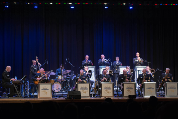 Eine Gruppe von Musikern auf einer Bühne, die Instrumente spielen, mit Mikrofonen und Ständern, Schildern mit der Aufschrift "The New Orleans Jazz Band", ein Vorhang im Hintergrund und ein Publikum darunter, das von Scheinwerfern beleuchtet wird.