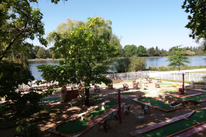Minigolf-Platz in einem Park mit grünem Gras, Bäumen und einem Zaun, umgeben von verstreuten Stühlen und Tischen, einem Gewässer mit einem Boot im Hintergrund und sichtbarem Himmel.