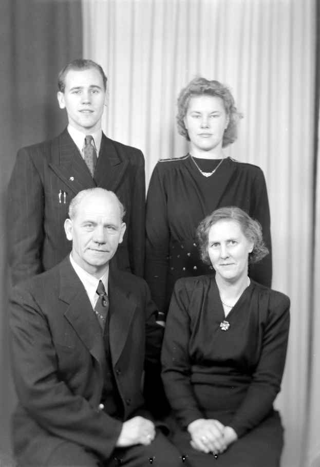 Ein Schwarz-Weiß-Foto von vier Personen, zwei Männern und zwei Frauen, die alle Anzüge tragen und vor einem Vorhang stehen.