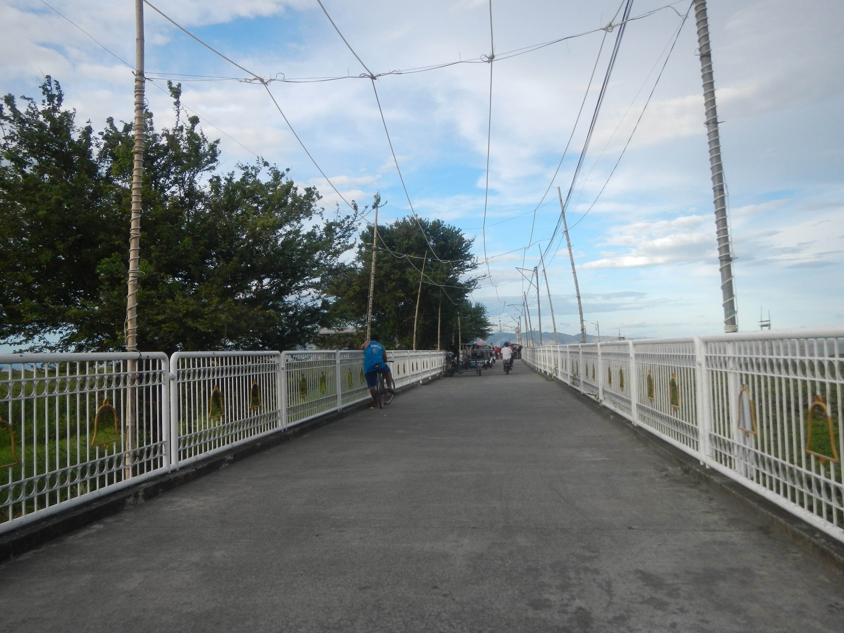 Eine Fußgängerbrücke mit Fahrradfahrern, Geländern, Strommasten mit Drähten, Bäumen und einem bewölkten Himmel im Hintergrund.