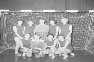 Schwarzes und weißes Foto einer Gruppe von Frauen, die vor einem Tor posieren, mit einem Fußball auf dem Boden und einem Vorhang im Hintergrund.