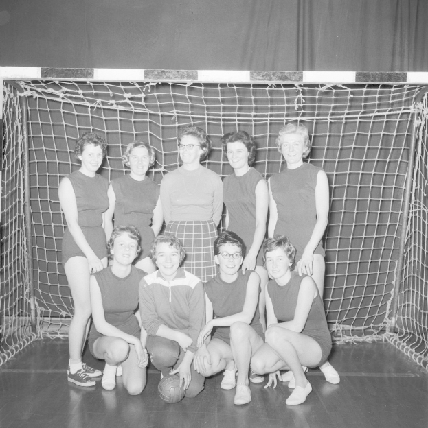 Schwarzes und weißes Foto einer Gruppe von Frauen, die vor einem Tor posieren, mit einem Fußball auf dem Boden und einem Vorhang im Hintergrund.