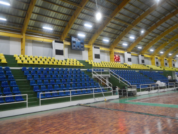 Großer Hallensportplatz mit Holzboden, blauen und gelben Sitzplätzen, Geländern, Stufen, wandmontierten Bildschirmen, Deckenleuchten und einigen Menschen.