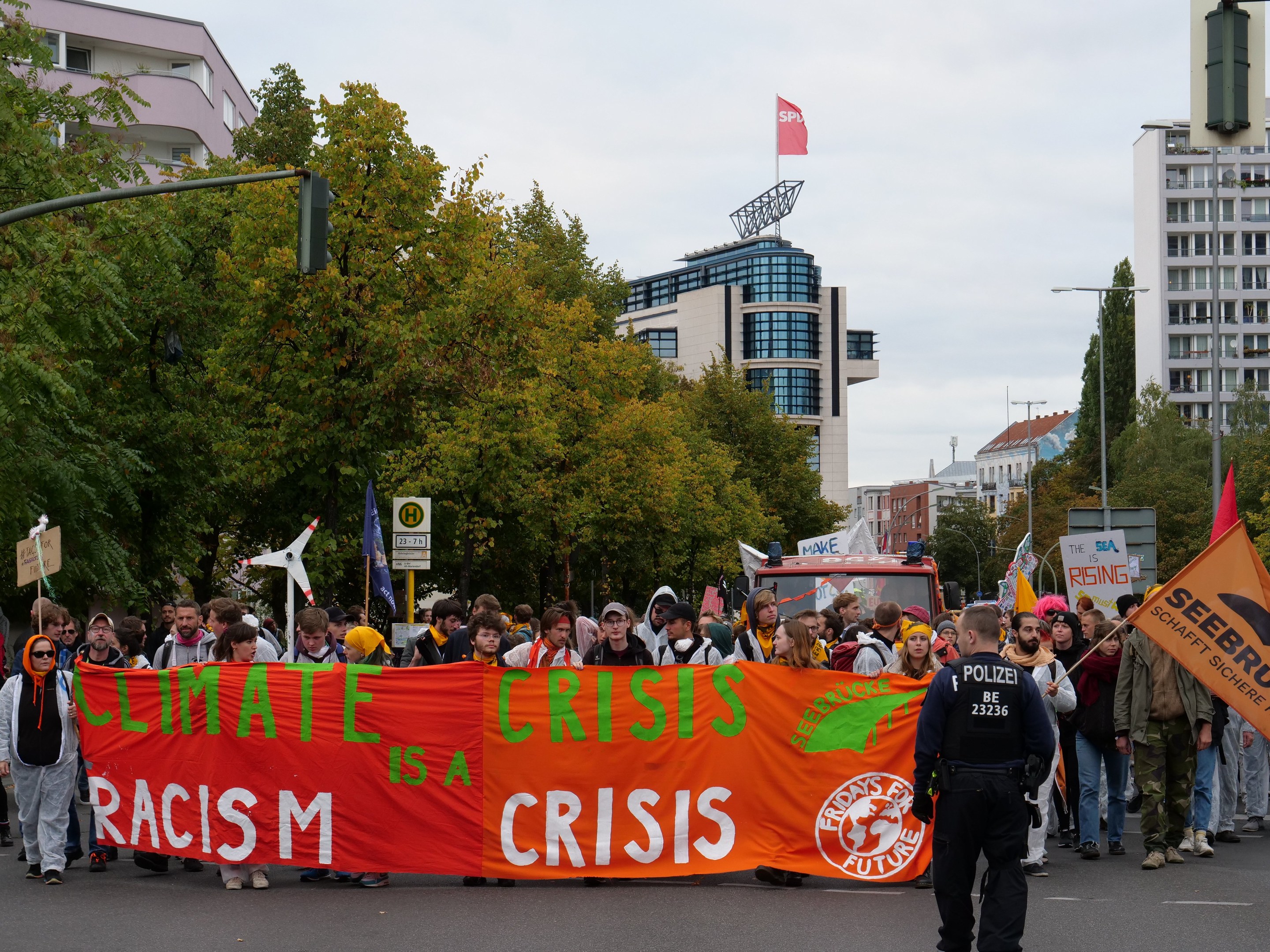 Menschen marschieren mit einem "Klimakrise ist eine Krise"-Schild die Straße entlang, mit Bäumen, Laternenmasten und geparkten Fahrzeugen an der Straße, Gebäude im Hintergrund bei einem klaren blauen Himmel.