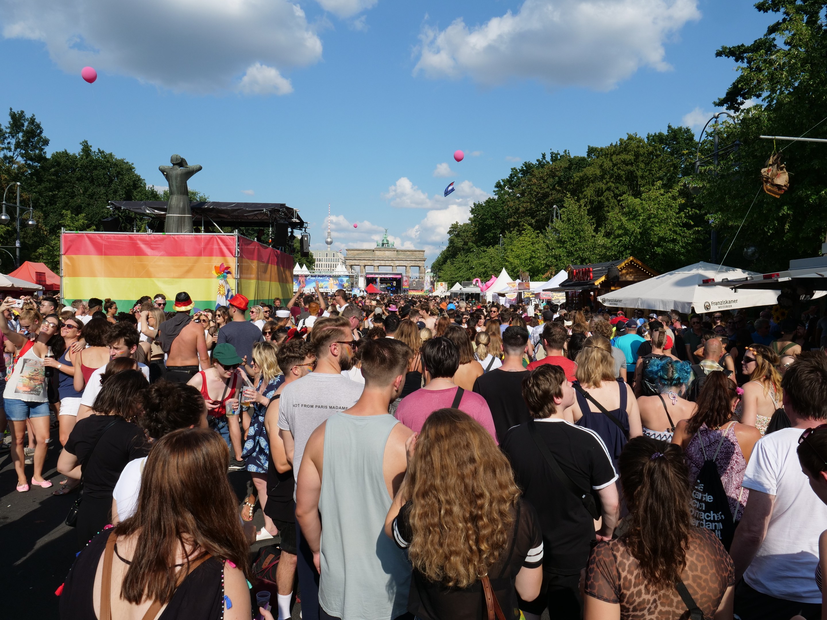 Eine gro├če Menge geht eine Stra├če entlang, die mit Zelten, B├Ąumen, Pfosten, Lichtern und einer Statue ges├Ąumt ist, w├Ąhrend des Berliner Pride-Festivals, mit Geb├Ąuden, Wolken und Ballons im Hintergrund.
