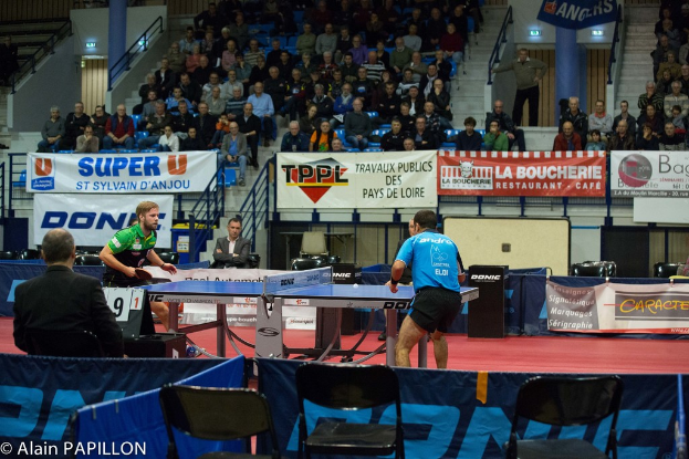 Zwei Männer spielen Tischtennis drinnen, mit mehreren Zuschauern, die stehen und sitzen, einigen leeren Stühlen und Werbetafeln.