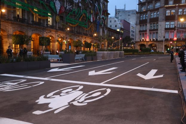 Eine Straße in einer Stadt mit einem Fahrradweg, Gebäuden, Straßenmasten, Straßenlaternen, Ampeln, Menschen auf dem Gehweg, Bänke, Pflanzen, Bäume, Schmuckstücke und einen klaren blauen Himmel.