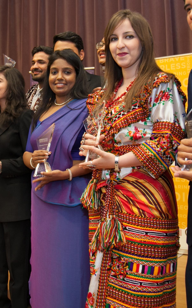 Eine Gruppe von Menschen auf einer Bühne mit einer Frau in der Mitte, die eine Trophäe hält, vor einem Vorhang und einer Tafel mit der Aufschrift "Bravo Blessing Award Gewinner."