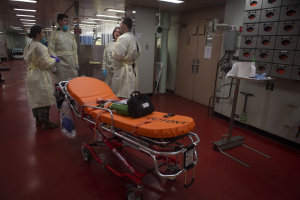 Eine Gruppe von Menschen in Schutzausrüstung um ein Bett in einem Krankenhauszimmer mit medizinischer Ausrüstung, einer Trage, einem Ständer, einer Tür, einer Säule, einem Schild und Deckenleuchten.