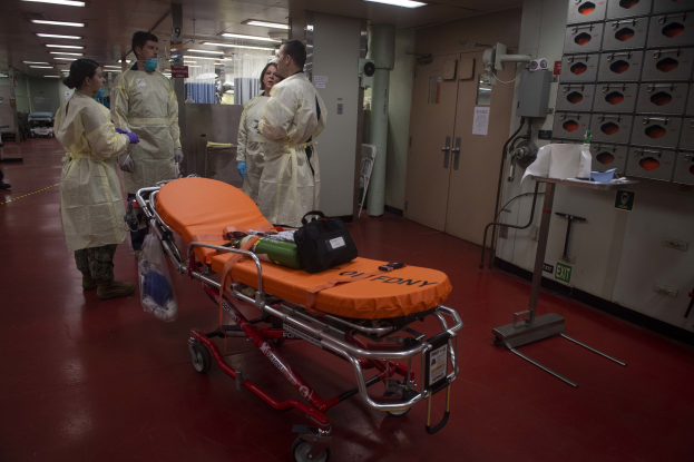 Eine Gruppe von Menschen in Schutzausrüstung um ein Bett in einem Krankenhauszimmer mit medizinischer Ausrüstung, einer Trage, einem Ständer, einer Tür, einer Säule, einem Schild und Deckenleuchten.