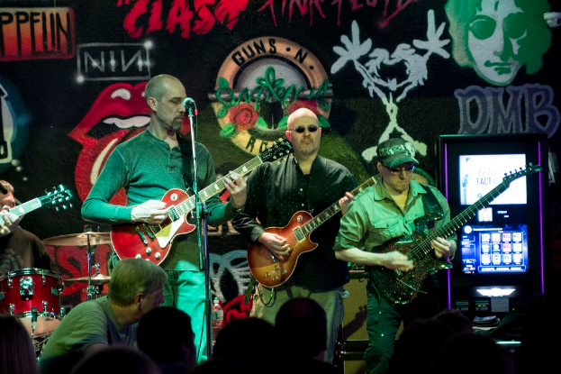 Drei Männer, die auf einer Bühne Gitarren spielen, vor einem Publikum und einer bemalten Wand.