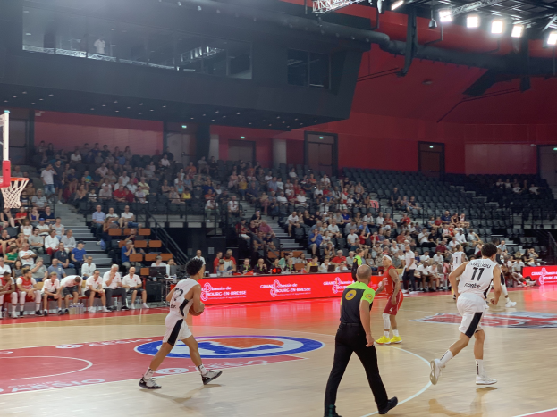 Eine Gruppe von Menschen, die Basketball in einer Turnhalle mit einem Netz auf der linken Seite und Zuschauern im Hintergrund spielen.
