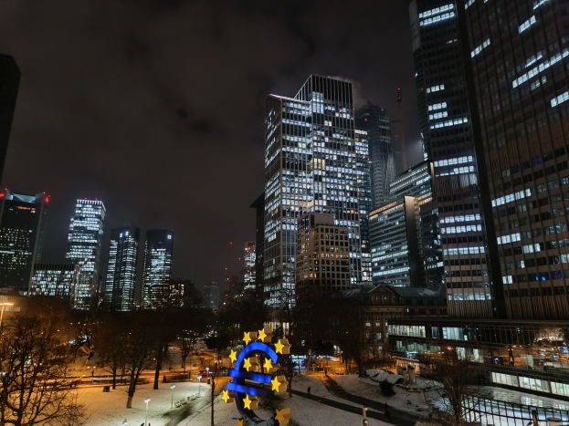 Europäische Zentralbank-Hauptquartier in Frankfurt, Deutschland bei Nacht mit beleuchteten Gebäuden, Bäumen, Straßenlaternen, Menschen und Wolken am Himmel.