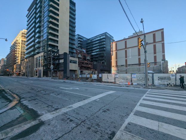 Eine Stadtstraße mit hohen Gebäuden, Straßenlaternen, Ampeln, Strommasten, Kabeln, Fahrzeugen, Fußgängern, Absperrpoller, Namenschilder, Bäumen und einem klaren blauen Himmel.