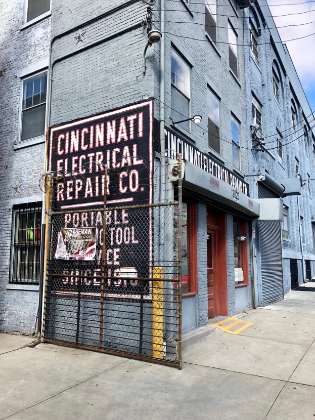 Das Gebäude der Cincinnati Electrical Repair Co. in Brooklyn, New York City, mit Fenstern, einer Tür, einem Schild, einem Metallzaun, einem Straßenpfahl und Drähten unter einem bewölktem Himmel.