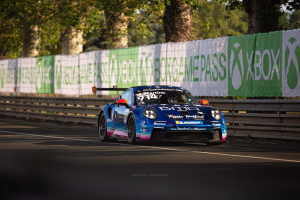 Blauer Porsche 911 GTS auf einer Rennstrecke mit einem Zaun und Banner im Hintergrund, Bäume und einen klaren blauen Himmel.
