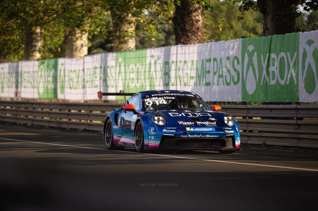 Blauer Porsche 911 GTS auf einer Rennstrecke mit einem Zaun und Banner im Hintergrund, Bäume und einen klaren blauen Himmel.