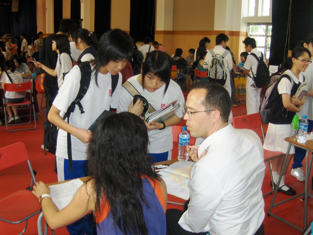 Ein Klassenzimmer mit mehreren Mädchen in weißen T-Shirts, die Bücher in den Händen halten, vorne, ein Mann und eine Frau im Vordergrund sitzend und diskutierend, und eine gelbe Wand mit einem Glasfenster im Hintergrund.