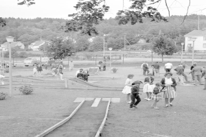 Schwarz-weißes Bild von Menschen, die auf einem Park mit Grünflächen, Laternen, geparkten Fahrzeugen und Gebäuden im Hintergrund Minigolf spielen.