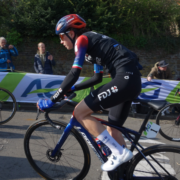Professionelle Radrennfahrerin Katie Archibald trägt Helm, Brille, Handschuhe und Schuhe und fährt ein Fahrrad auf einer Straße neben einer Menge, mit einem Banner, einer Wand, Bäumen und einem Gebäude im Hintergrund.