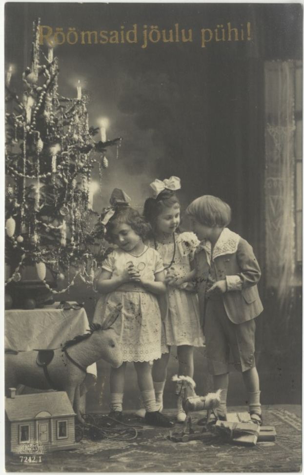 Ein altes Foto von drei Kindern, die vor einem geschmützten Weihnachtsbaum stehen, mit einem Tisch auf der linken Seite und verstreuten Spielsachen auf der rechten Seite und Text oben.