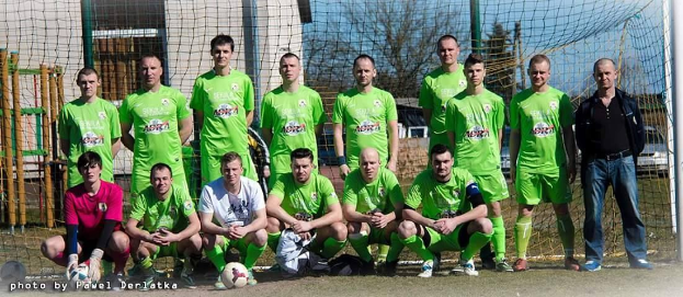 Eine Gruppe von Männern in grünen T-Shirts steht auf einem Fußballfeld mit einem Ball vor ihnen, in der Nähe eines Torpfostens, mit Bäumen, Gebäuden und Fahrzeugen im Hintergrund.