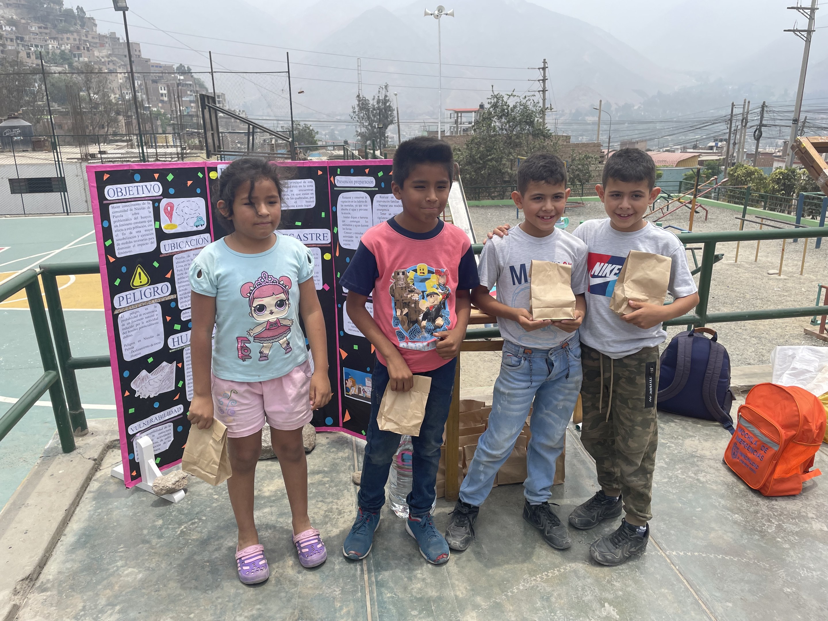 Gruppe von Kindern auf einem Basketballfeld mit Papiertüten in den Händen, mit Vorräten auf dem Boden, einer Tafel mit Text links daneben und Zäunen, Pfählen und Gebäuden im Hintergrund.