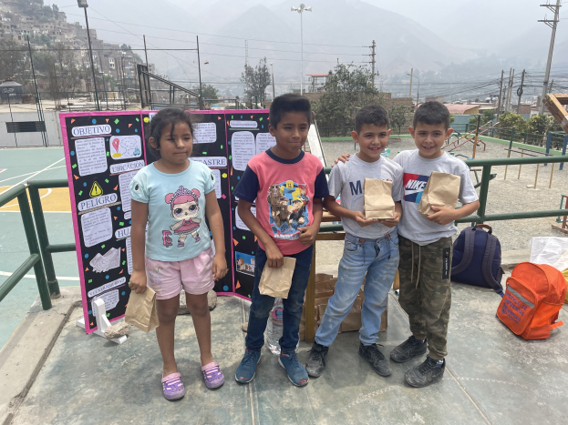 Gruppe von Kindern auf einem Basketballfeld mit Papiertüten in den Händen, mit Vorräten auf dem Boden, einer Tafel mit Text links daneben und Zäunen, Pfählen und Gebäuden im Hintergrund.