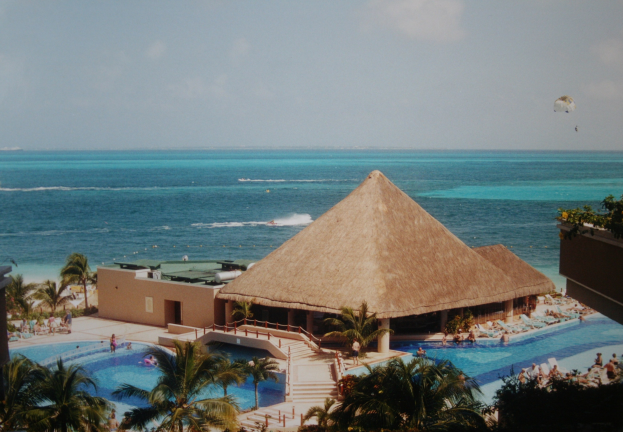 Resort mit einem gro├čen Schwimmbad umgeben von Palmen, Hütten und Menschen, überblickend einen Ozean unter einem bew├Âlktem Himmel mit einem Fallschirm in der Luft.