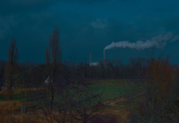 Outdoorszene mit bewölktem Himmel, einer rauchenden Fabrik, ein paar Häusern und einer grasbewachsenen Fläche, die mit Bäumen und Pflanzen gefüllt ist.
