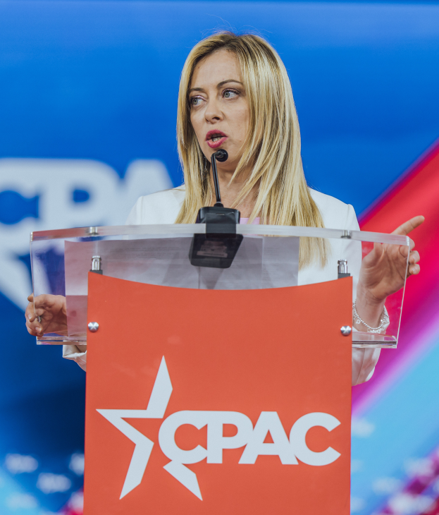 Eine Frau in einem weißen Kleid steht an einem Podium mit einem Mikrofon, unscharfer Hintergrund legt den Fokus auf sie, wahrscheinlich eine Politologin.