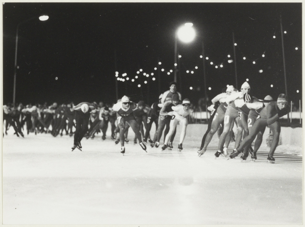Eine Gruppe von Menschen, die nachts auf einer Eisbahn Schlittschuh laufen, beleuchtet von Straßenlaternen im Hintergrund, in einem Schwarz-Weiß-Foto.