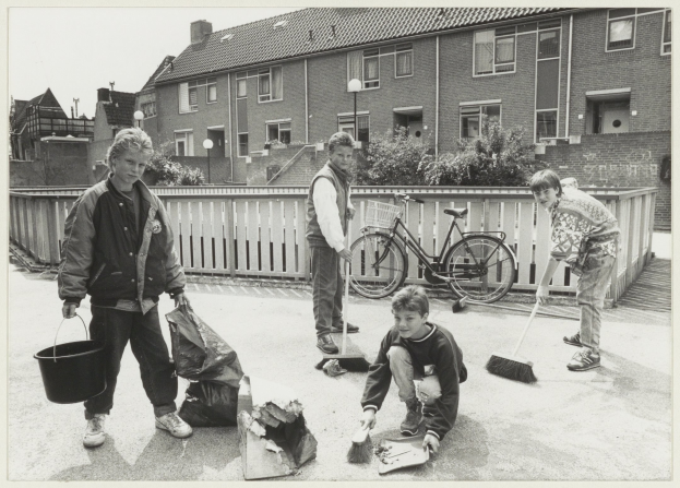 Vier Personen, zwei Jungen und zwei Mädchen, kehren eine Straße mit Besen, einer Junge hält einen Eimer und Deckel, mit einem Fahrrad, Zaun, Pflanzen, Laternenpfähle, Gebäude und einen klaren Himmel im Hintergrund.