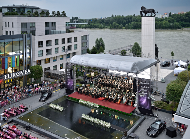 Eine große Menschenmenge sitzt auf einer Bühne neben einem Gewässer auf der Frankfurter Motor Show, einige halten Musikinstrumente, Fahrzeuge auf der Straße darunter, Banner mit Text, ein Dach mit einem Dach, eine Statue auf einem Pfeiler, Gebäude mit Fenstern, Bäume und Laternen im Hintergrund unter einem bewölkten Himmel.