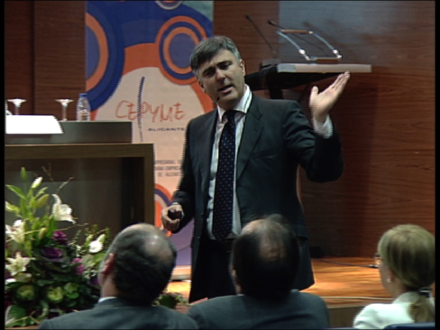 Ein Mann in einem schwarzen Blazer steht an einem Podium mit Mikrofonen, spricht in eines hinein, mit einer Blumenanordnung und einem Tisch mit einem Buch und einer Wasserflasche daneben, während drei Personen auf Stühlen davor sitzen, vor einer braunen Wand und einem bunten Schild mit Text.