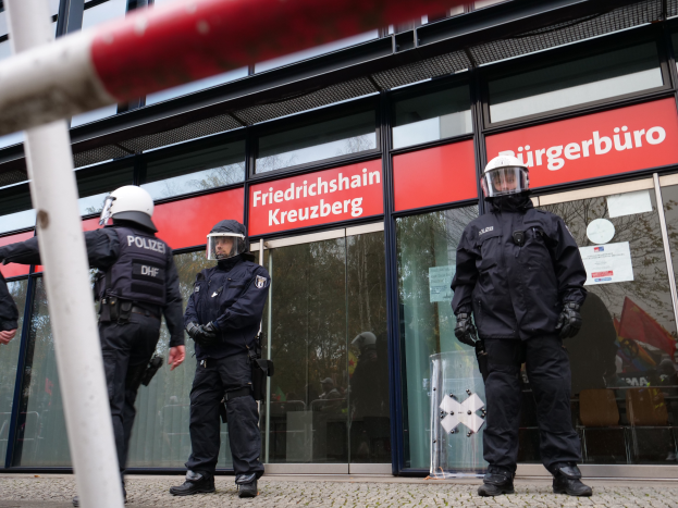 Eine Gruppe von Polizisten in Helmen und Handschuhen steht vor einem Glasgebäude mit einer Stange links und Schildern im Hintergrund, die "Friedrichshain Kreuzberg" und Papiere an den Glaswänden angeklebt zeigen.